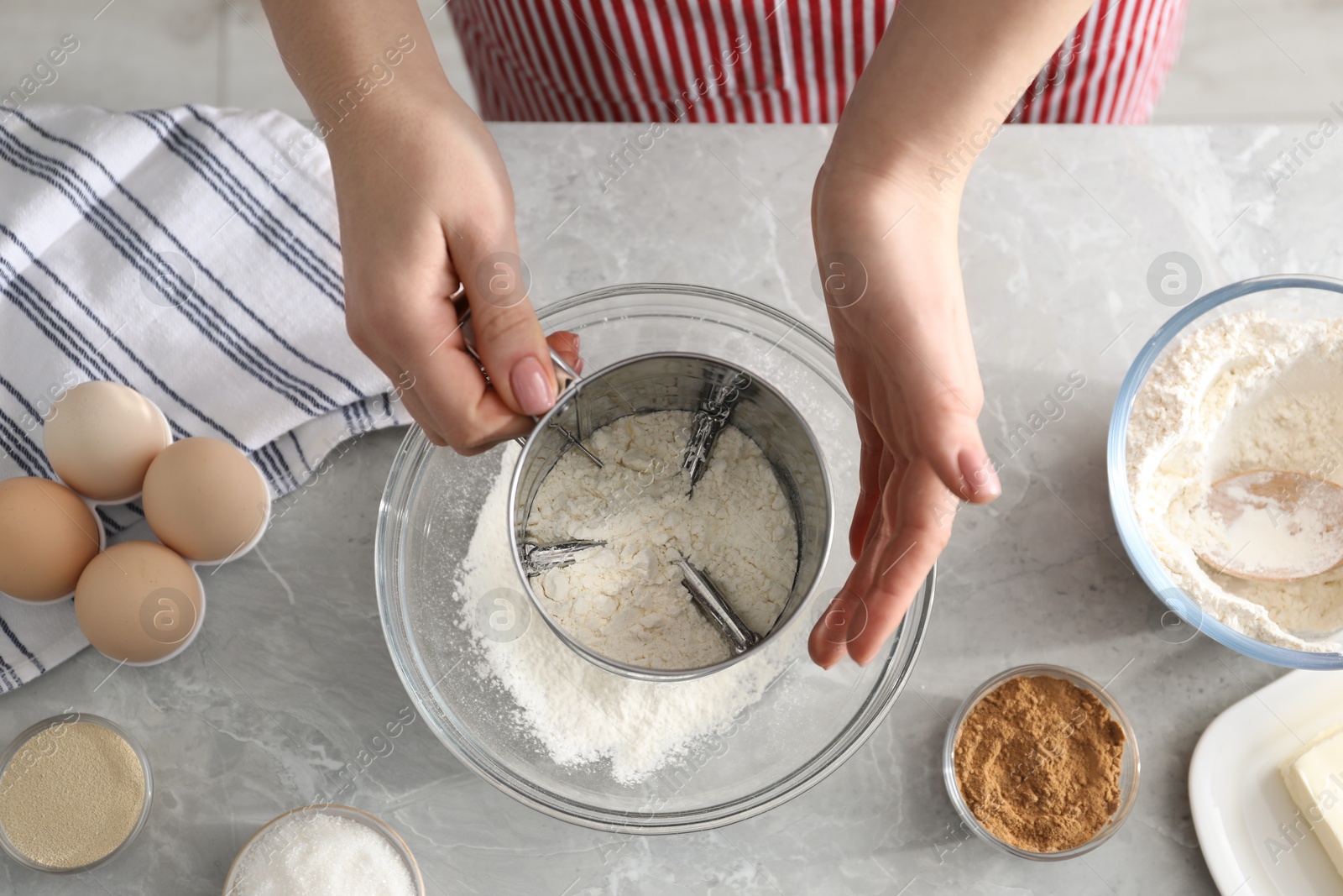 Photo of Making cinnamon rolls. Woman sieving flour at gray marble table, top view