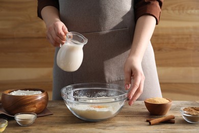 Photo of Making cinnamon rolls. Woman pouring milk into bowl at wooden table, closeup