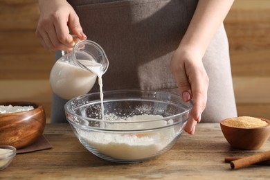 Photo of Making cinnamon rolls. Woman pouring milk into bowl at wooden table, closeup