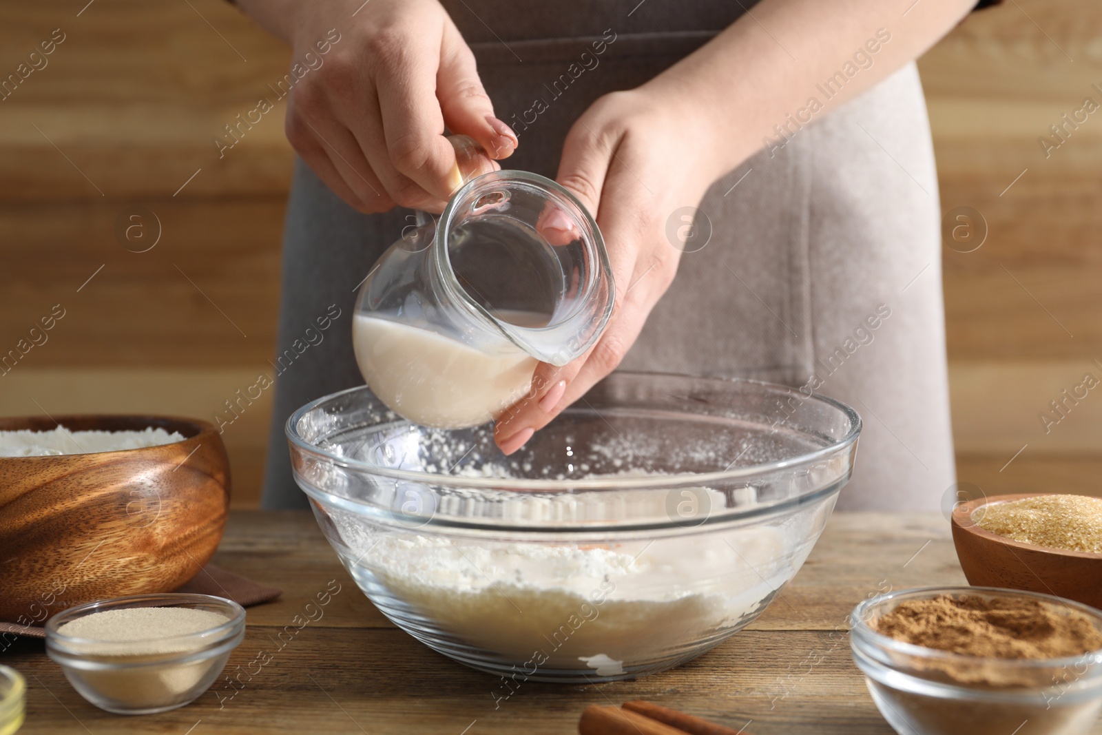 Photo of Making cinnamon rolls. Woman pouring milk into bowl at wooden table, closeup