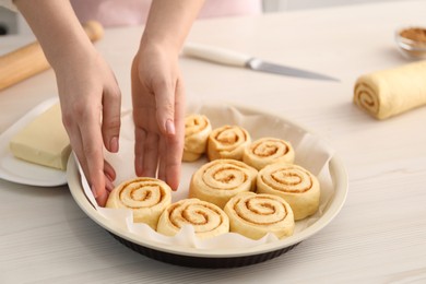 Photo of Woman putting cinnamon roll into baking dish at white wooden table, closeup