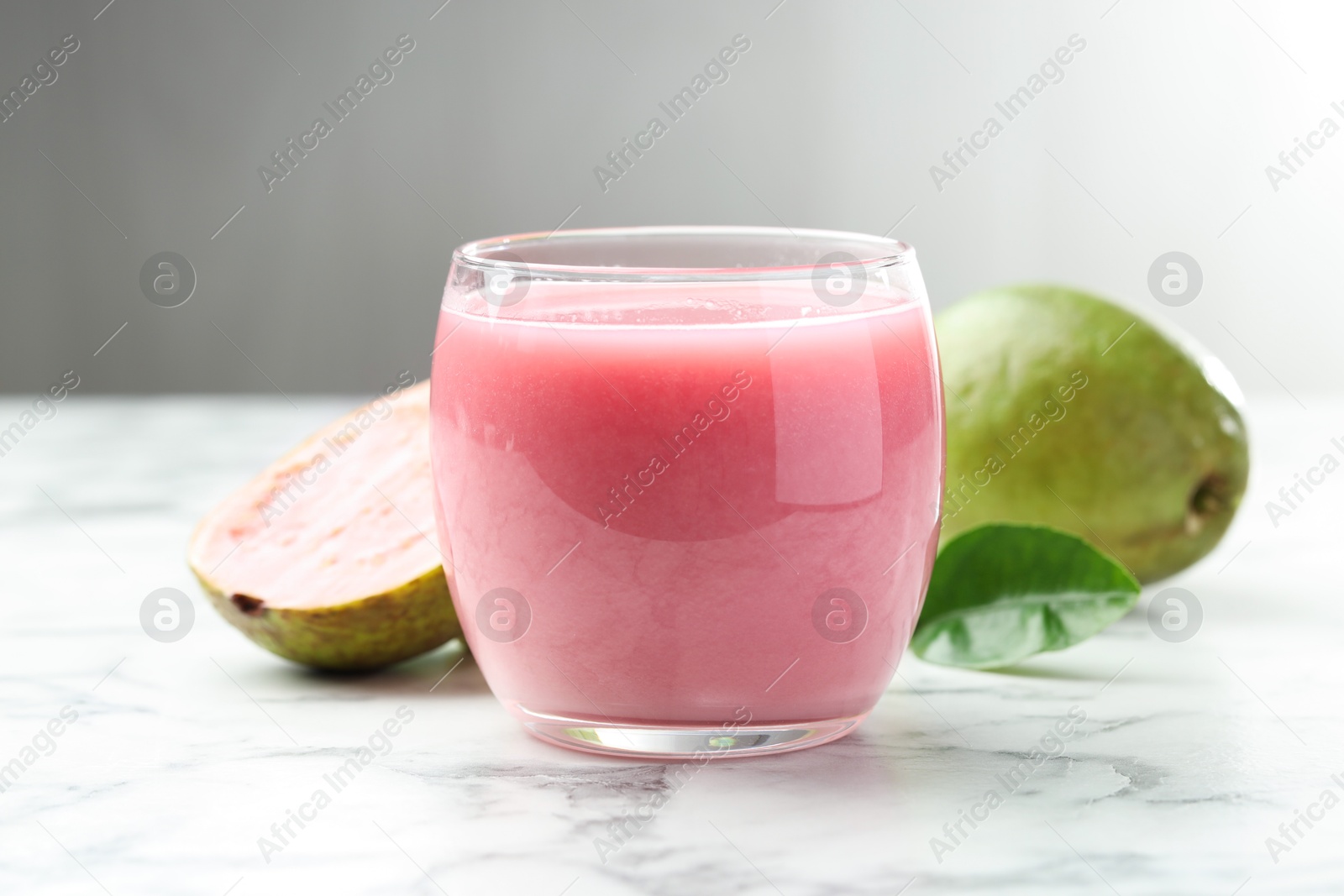 Photo of Tasty guava juice in glass, leaf and fruits on white marble table against grey background, closeup