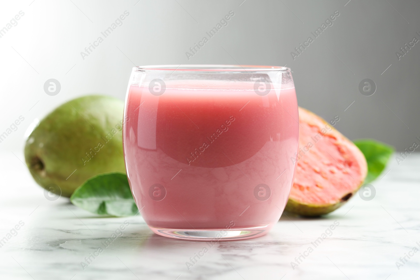 Photo of Tasty guava juice in glass, leaves and fruits on white marble table against grey background, closeup