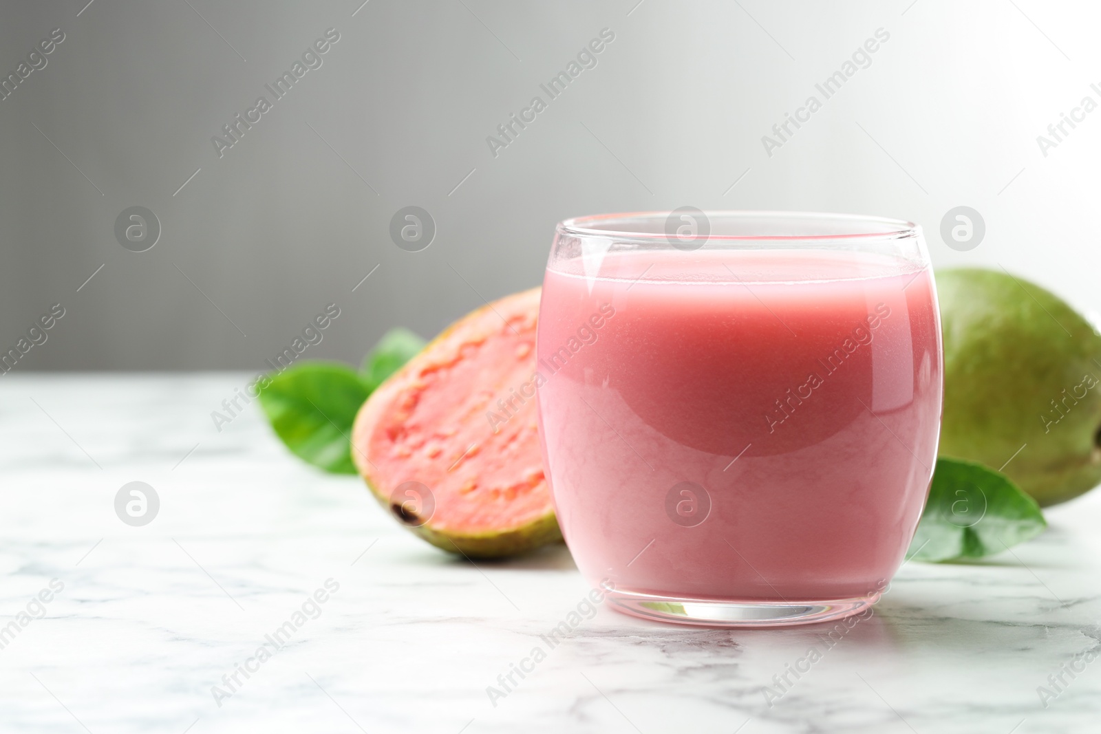 Photo of Tasty guava juice in glass, leaves and fruits on white marble table against grey background, closeup. Space for text