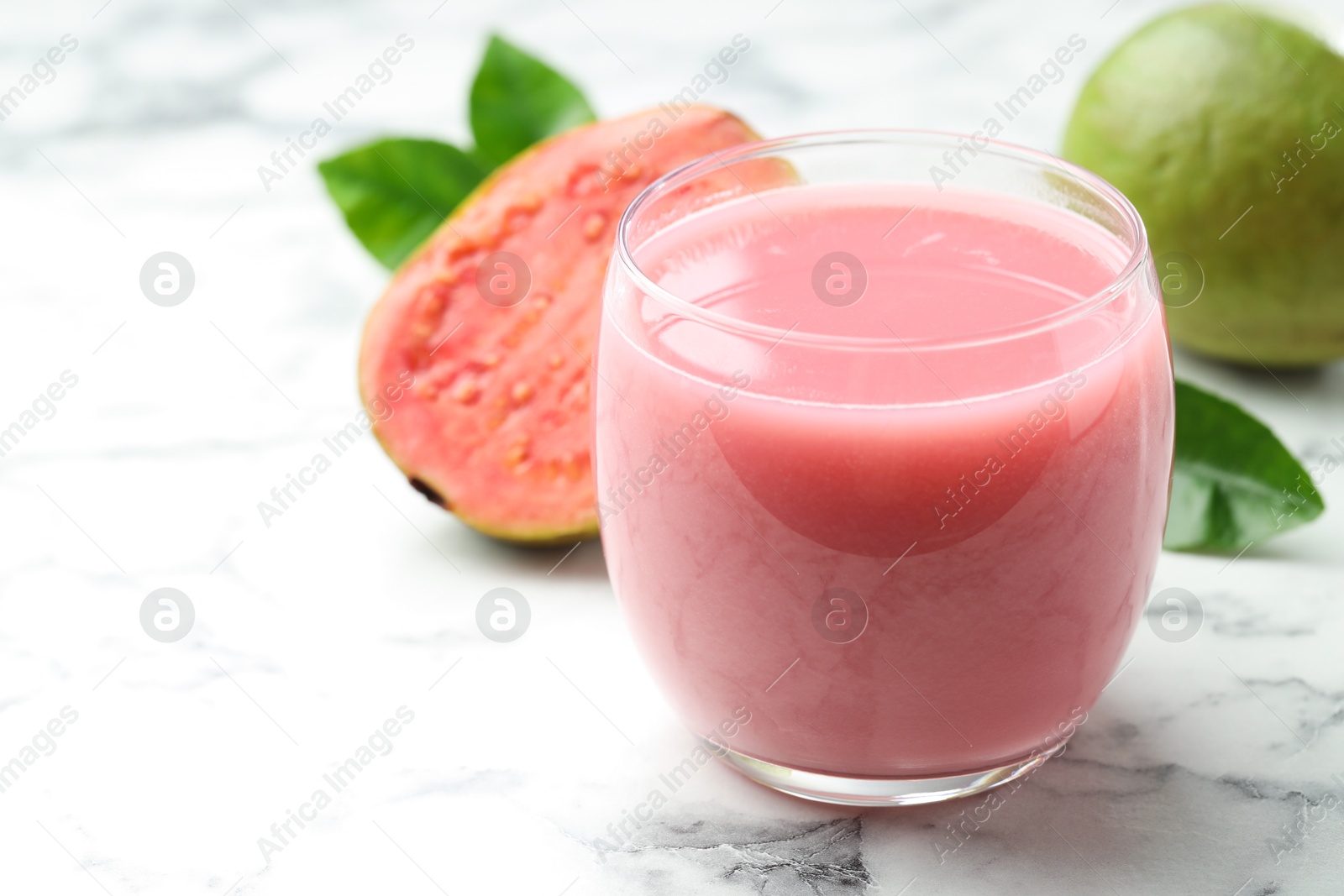 Photo of Tasty guava juice in glass, leaves and fruits on white marble table, closeup. Space for text