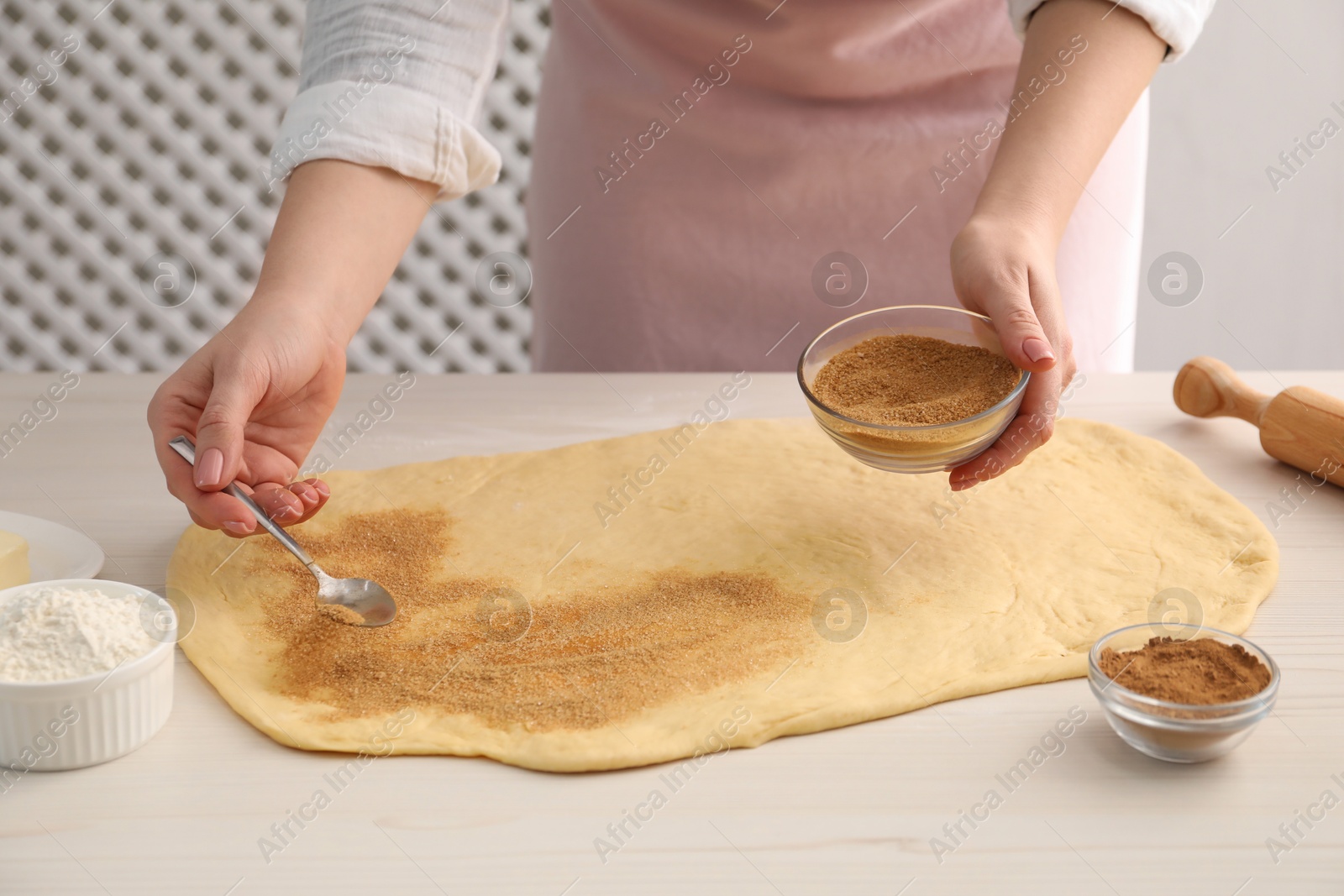 Photo of Making cinnamon rolls. Woman adding spice with sugar into dough at white wooden table, closeup