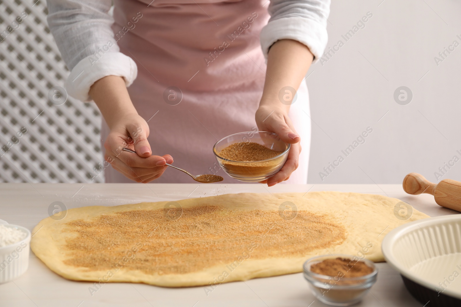 Photo of Making cinnamon rolls. Woman adding spice with sugar into dough at white wooden table, closeup