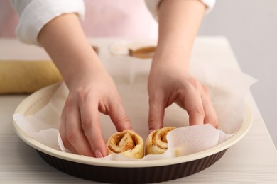 Photo of Woman putting cinnamon roll into baking dish at white wooden table, closeup