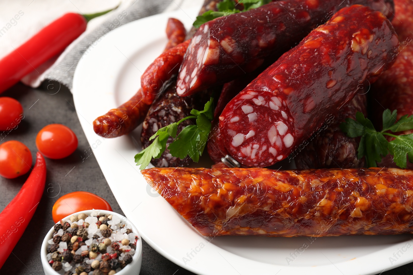 Photo of Different smoked sausages, parsley, peppercorns, tomatoes and chili pepper on grey textured table, closeup