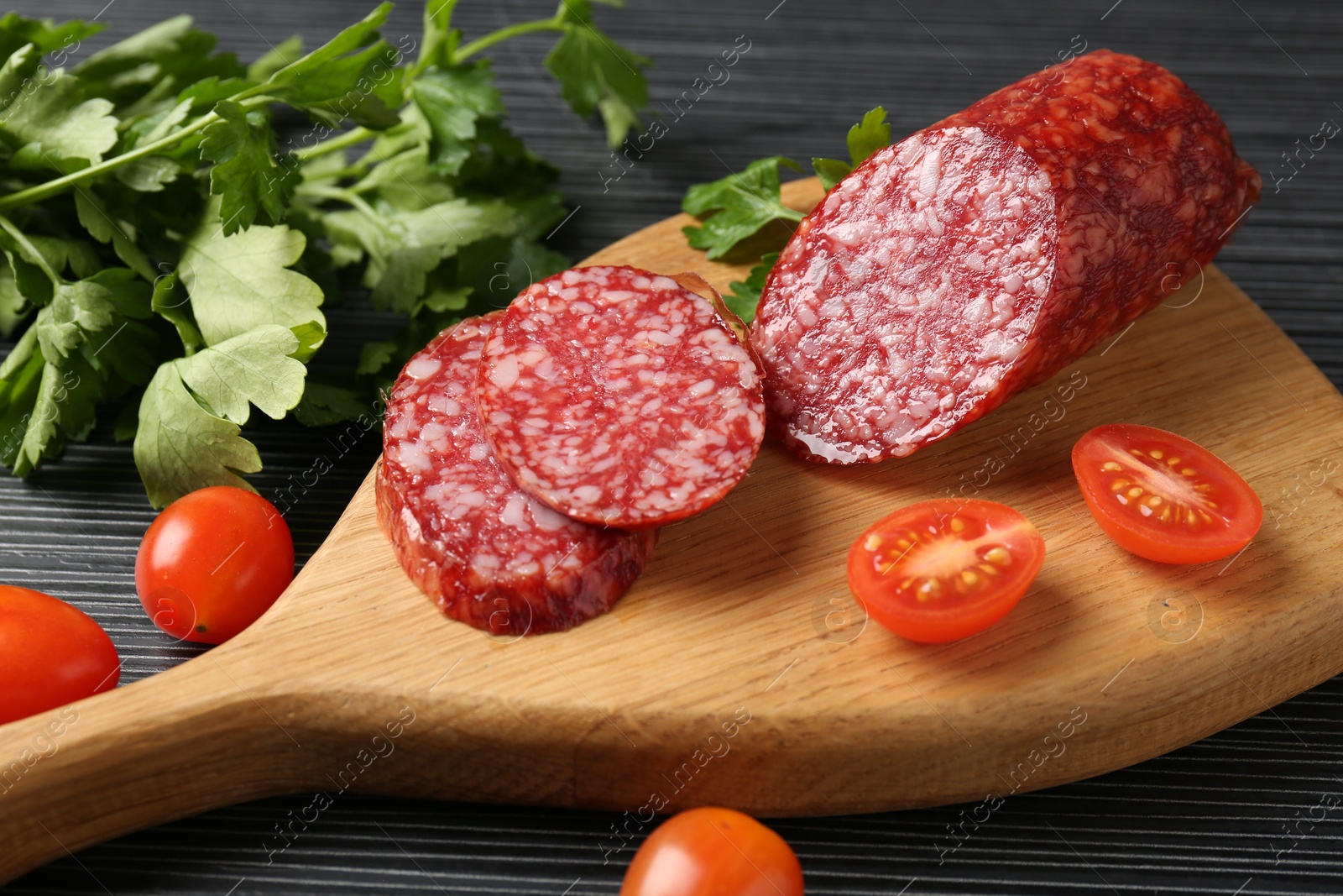 Photo of Cut smoked sausage, tomatoes and parsley on black wooden table, closeup