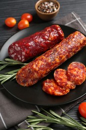 Photo of Cut smoked sausages, rosemary and tomatoes on black wooden table, closeup