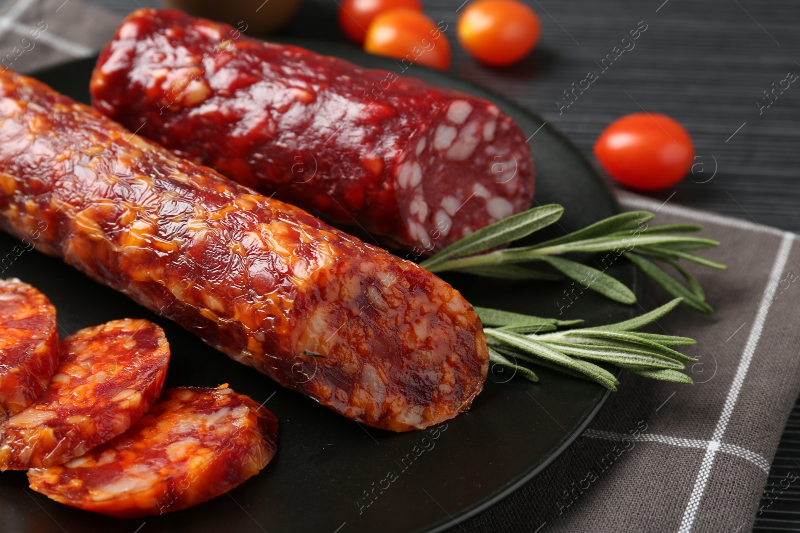 Photo of Cut smoked sausages, rosemary and tomatoes on black wooden table, closeup