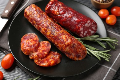 Photo of Cut smoked sausages, rosemary and tomatoes on black wooden table, closeup