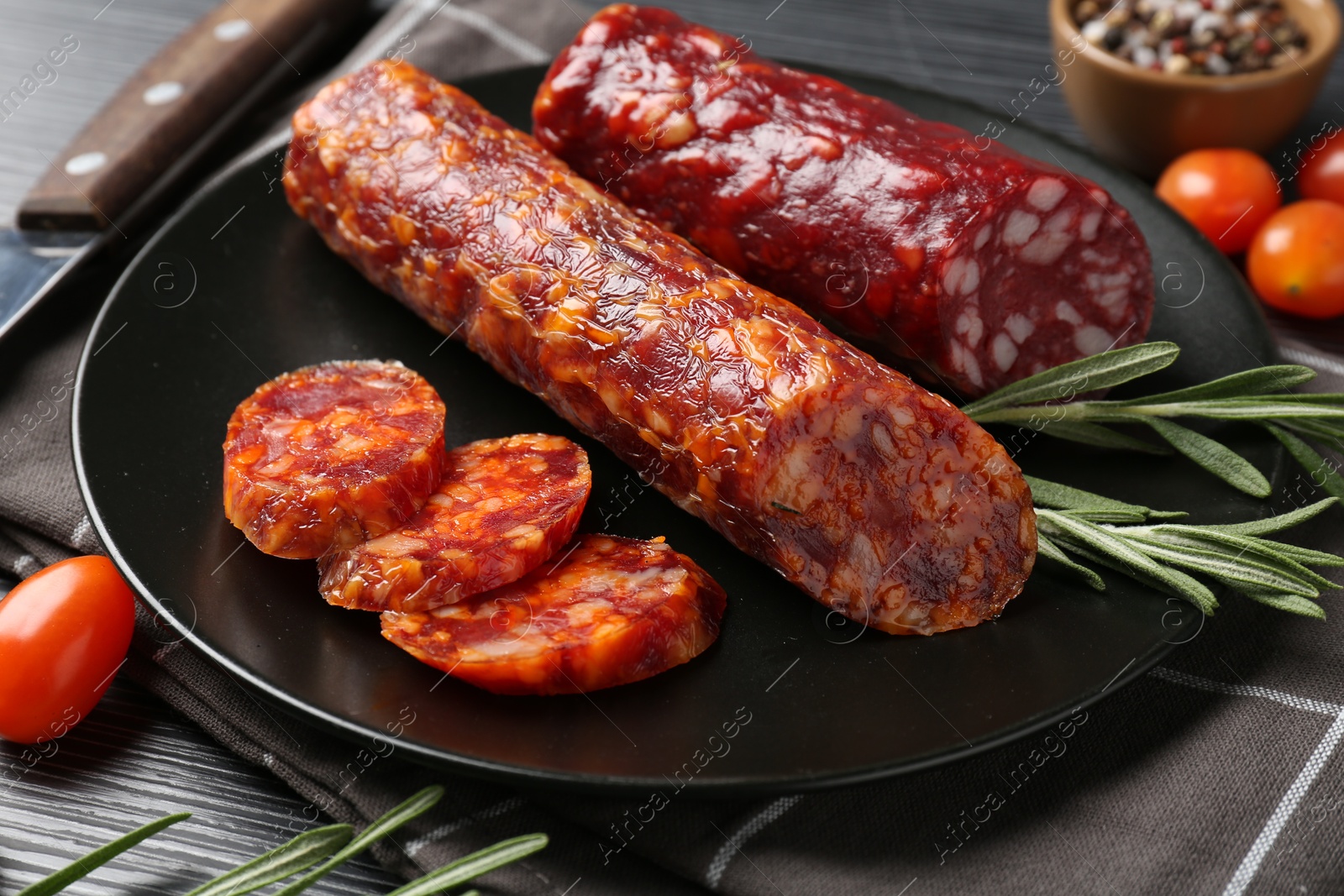 Photo of Cut smoked sausages, rosemary and tomatoes on black wooden table, closeup