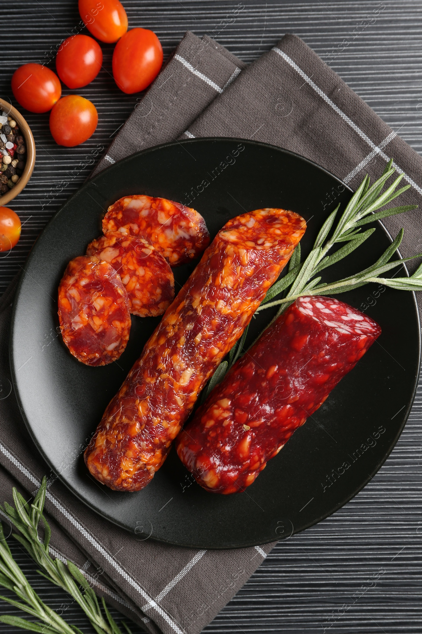 Photo of Cut smoked sausages, rosemary and tomatoes on black wooden table, flat lay