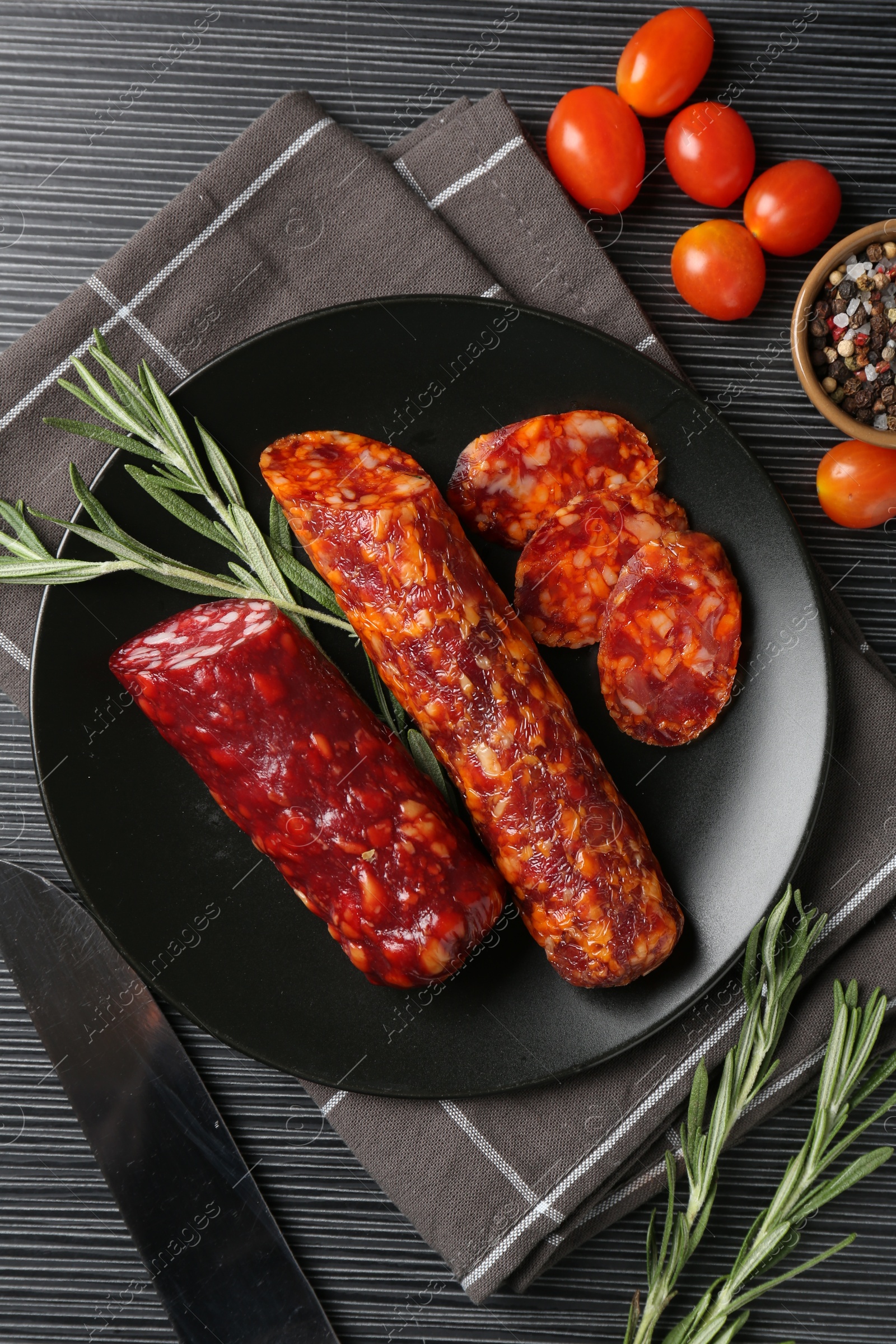 Photo of Cut smoked sausages, rosemary and tomatoes on black wooden table, flat lay