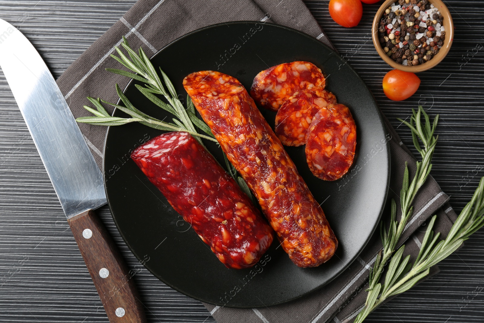 Photo of Cut smoked sausages, rosemary, peppercorns and tomatoes on black wooden table, flat lay