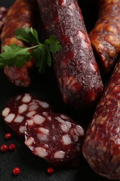 Photo of Different smoked sausages, parsley and peppercorns on black table, closeup