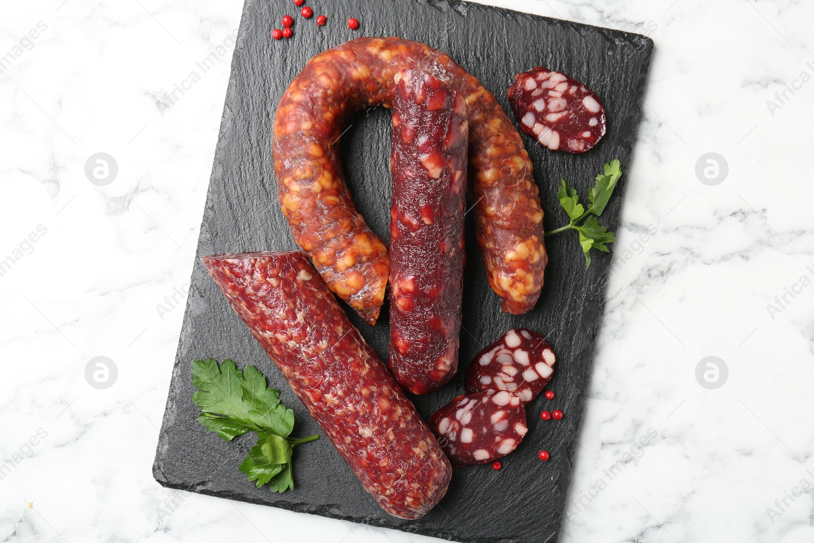 Photo of Different smoked sausages, parsley and peppercorns on white marble table, top view