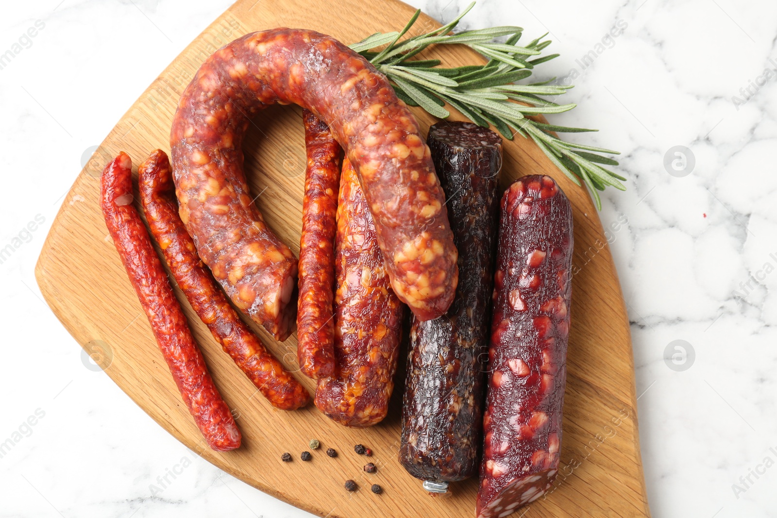 Photo of Different smoked sausages, rosemary and peppercorns on white marble table, top view