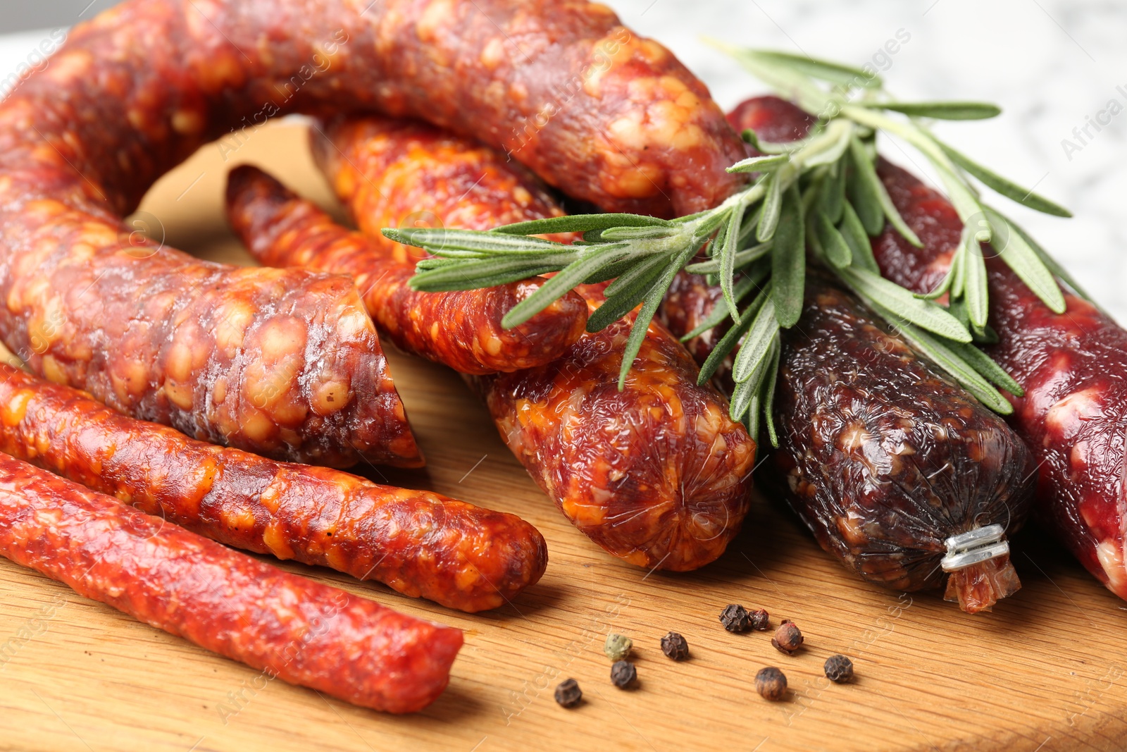 Photo of Different smoked sausages, rosemary and peppercorns on wooden board, closeup