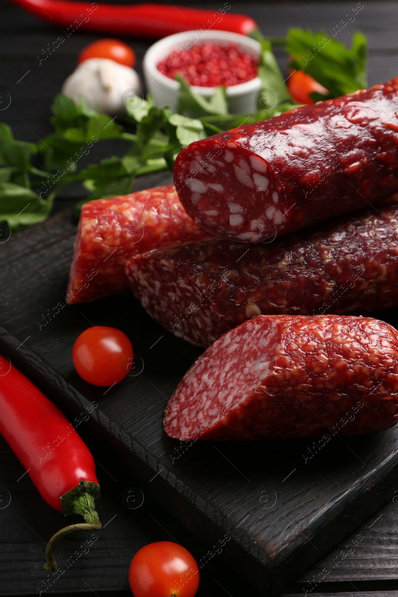 Photo of Different smoked sausages, tomatoes and chili pepper on black wooden table, closeup