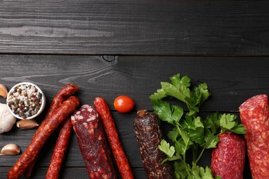 Photo of Different smoked sausages, tomato, parsley and peppercorns on black wooden table, flat lay. Space for text