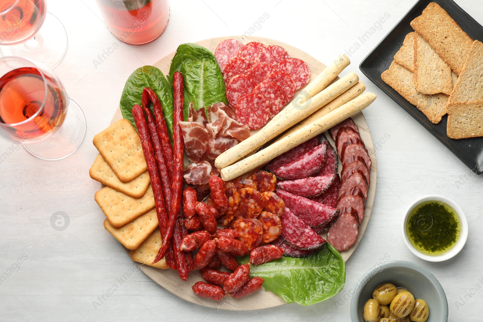 Photo of Different smoked sausages and other snacks served with wine on white table, flat lay