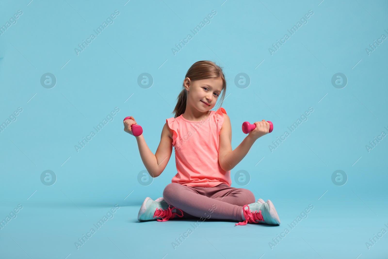 Photo of Cute little girl exercising with dumbbells on light blue background