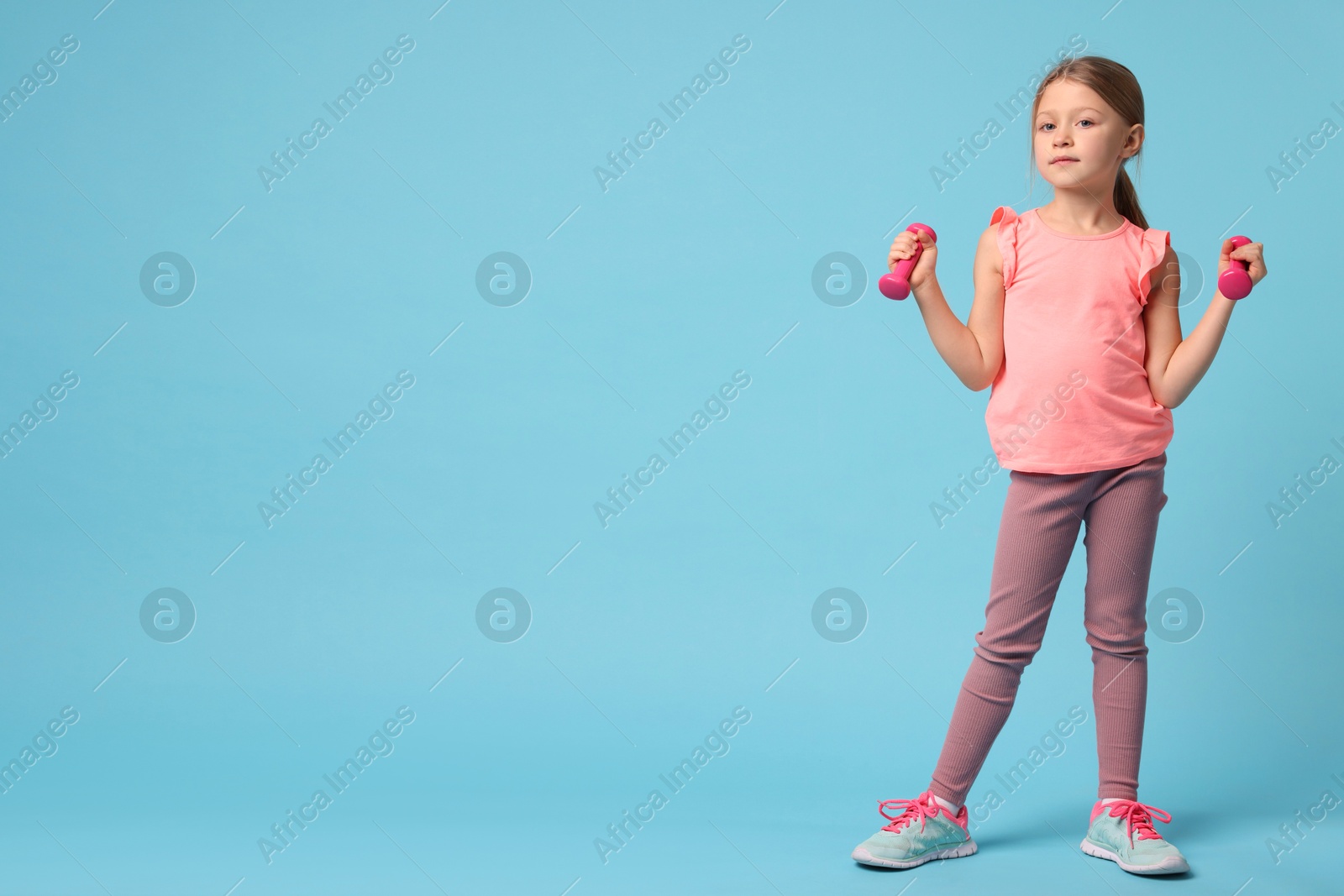 Photo of Cute little girl exercising with dumbbells on light blue background, space for text