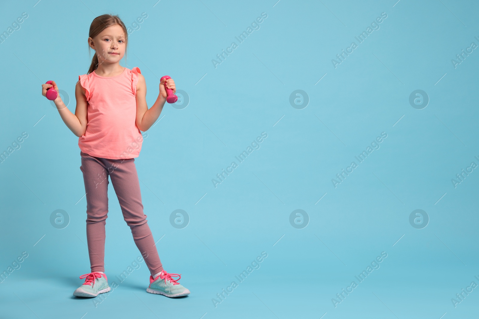 Photo of Cute little girl exercising with dumbbells on light blue background, space for text