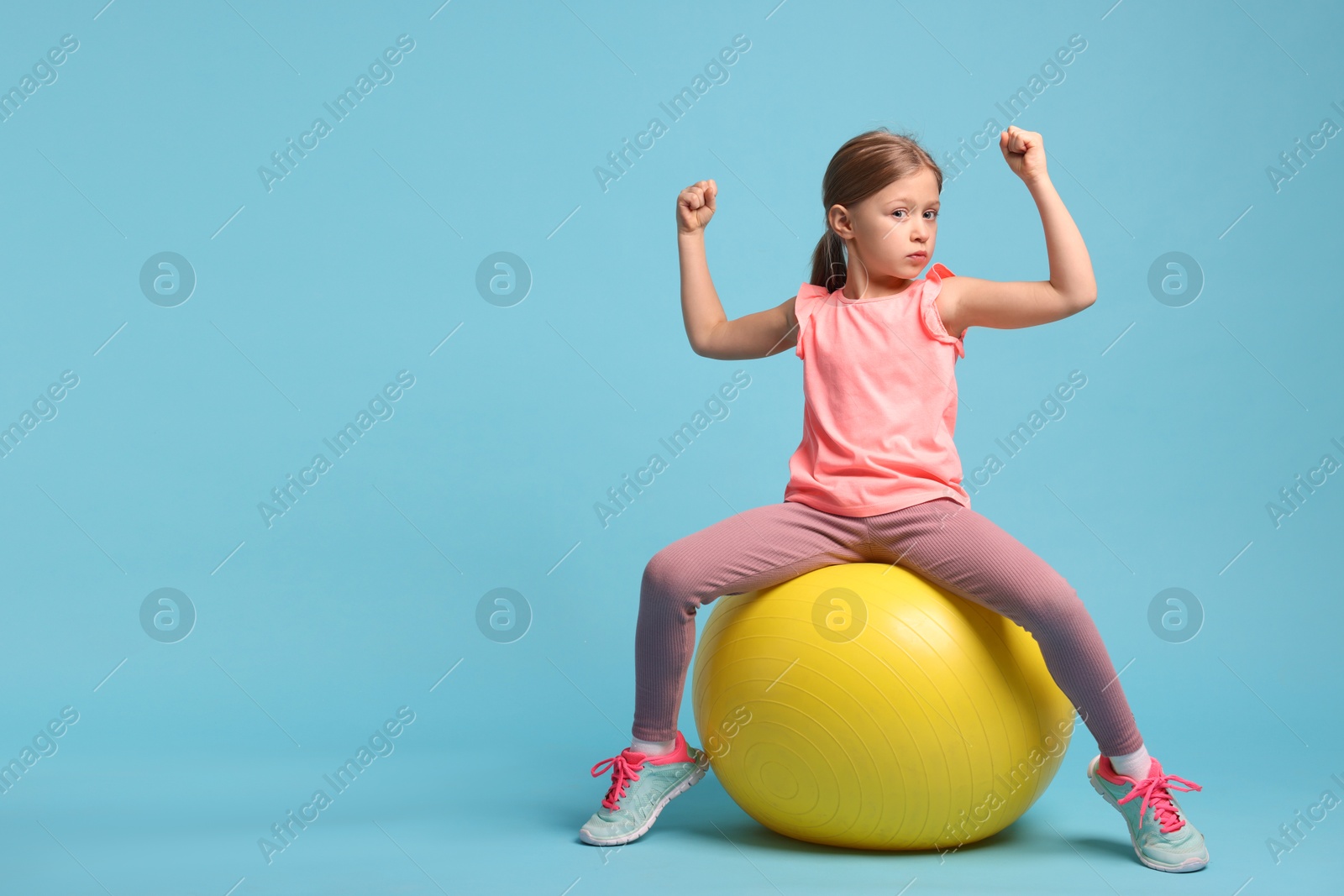 Photo of Cute little girl with fitness ball showing her biceps on light blue background, space for text