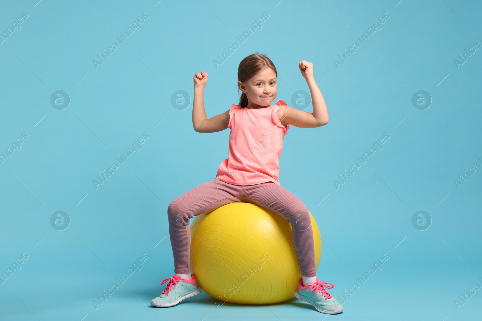 Photo of Cute little girl with fitness ball showing her biceps on light blue background