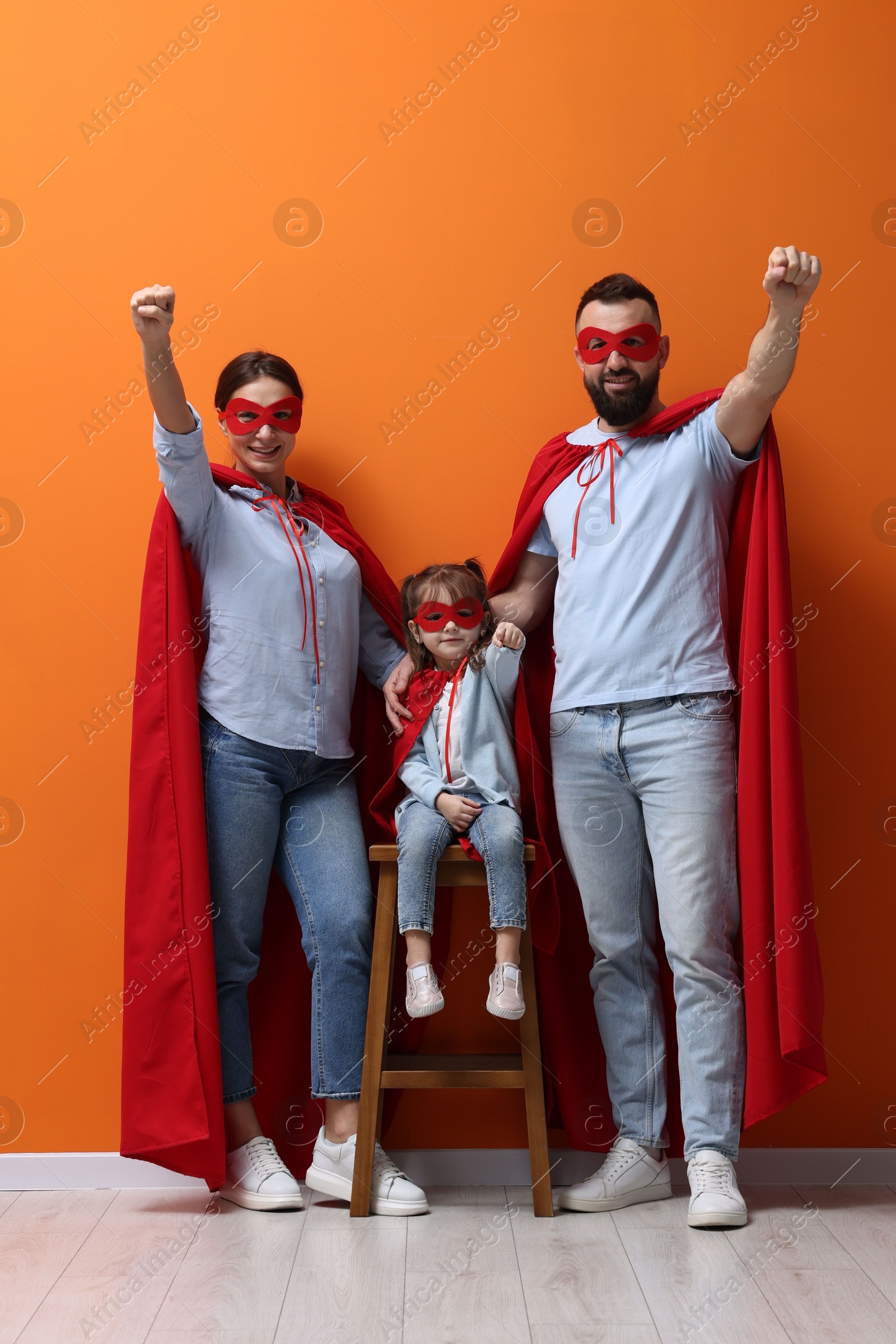 Photo of Parents and their cute little daughter wearing superhero costumes near orange wall indoors