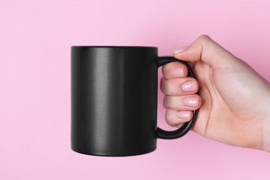 Photo of Woman holding blank black mug on pink background, closeup. Mockup for design