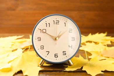 Photo of Autumn time. Alarm clock among leaves on wooden table, closeup