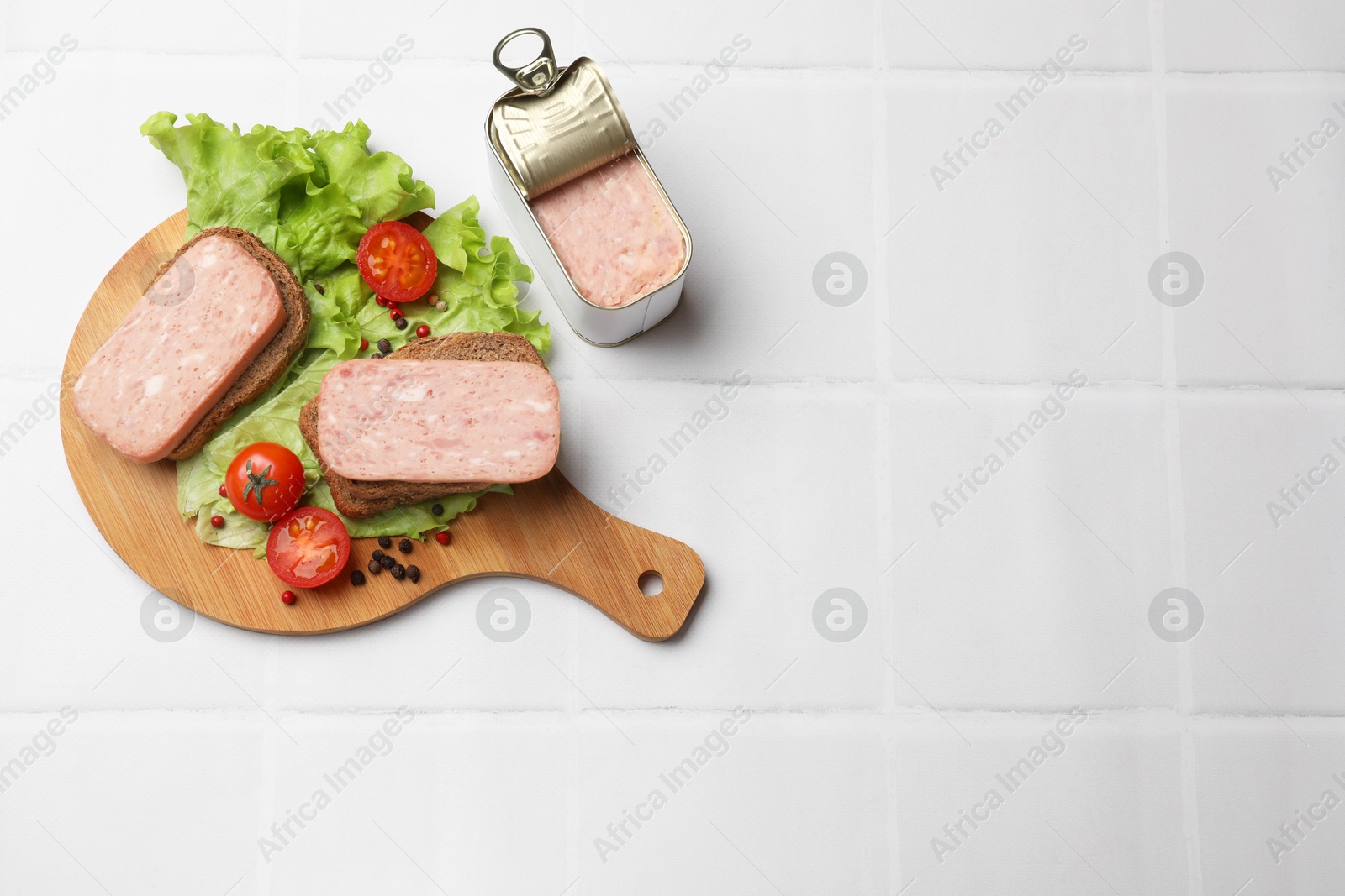 Photo of Tasty canned meat, bread, tomatoes, spices and lettuce on white tiled table, top view. Space for text