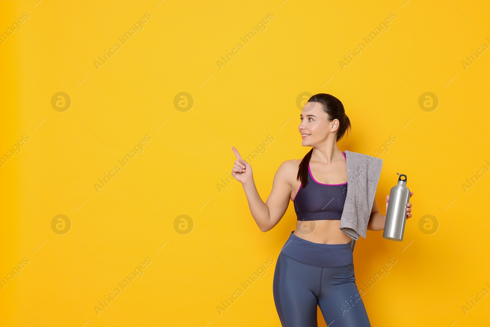 Photo of Smiling woman with thermo bottle and towel pointing at something on yellow background. Space for text