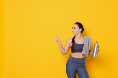 Photo of Smiling woman with thermo bottle and towel pointing at something on yellow background. Space for text