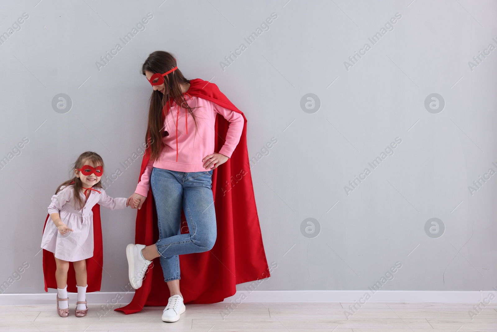 Photo of Mother and her cute little daughter wearing superhero costumes near gray wall indoors, space for text