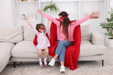 Photo of Mother and her cute little daughter wearing superhero costumes on sofa at home