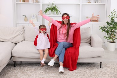 Photo of Mother and her cute little daughter wearing superhero costumes on sofa at home