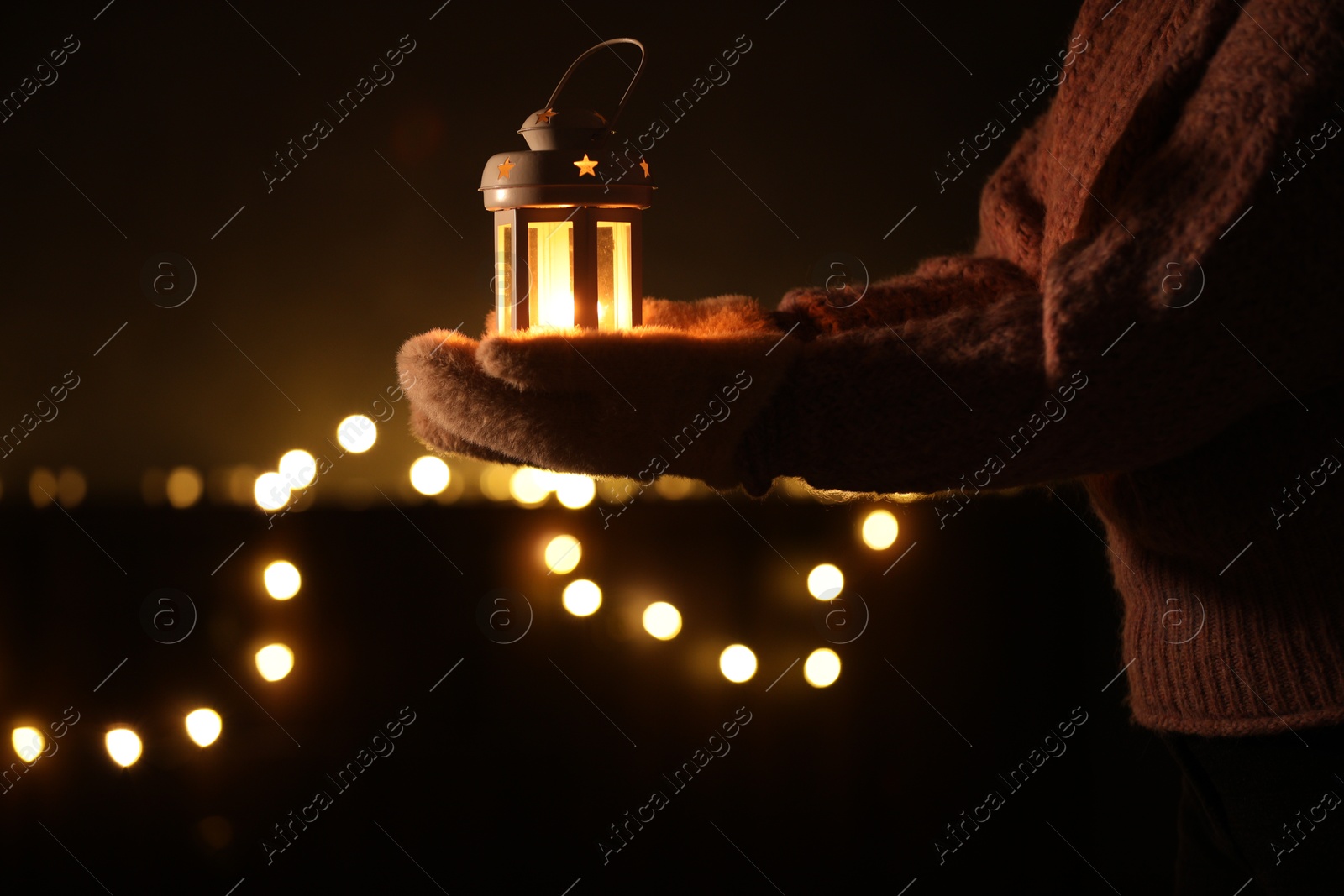 Photo of Woman holding Christmas lantern with burning candle in darkness, closeup. Bokeh effect