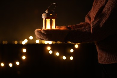 Photo of Woman holding Christmas lantern with burning candle in darkness, closeup. Bokeh effect