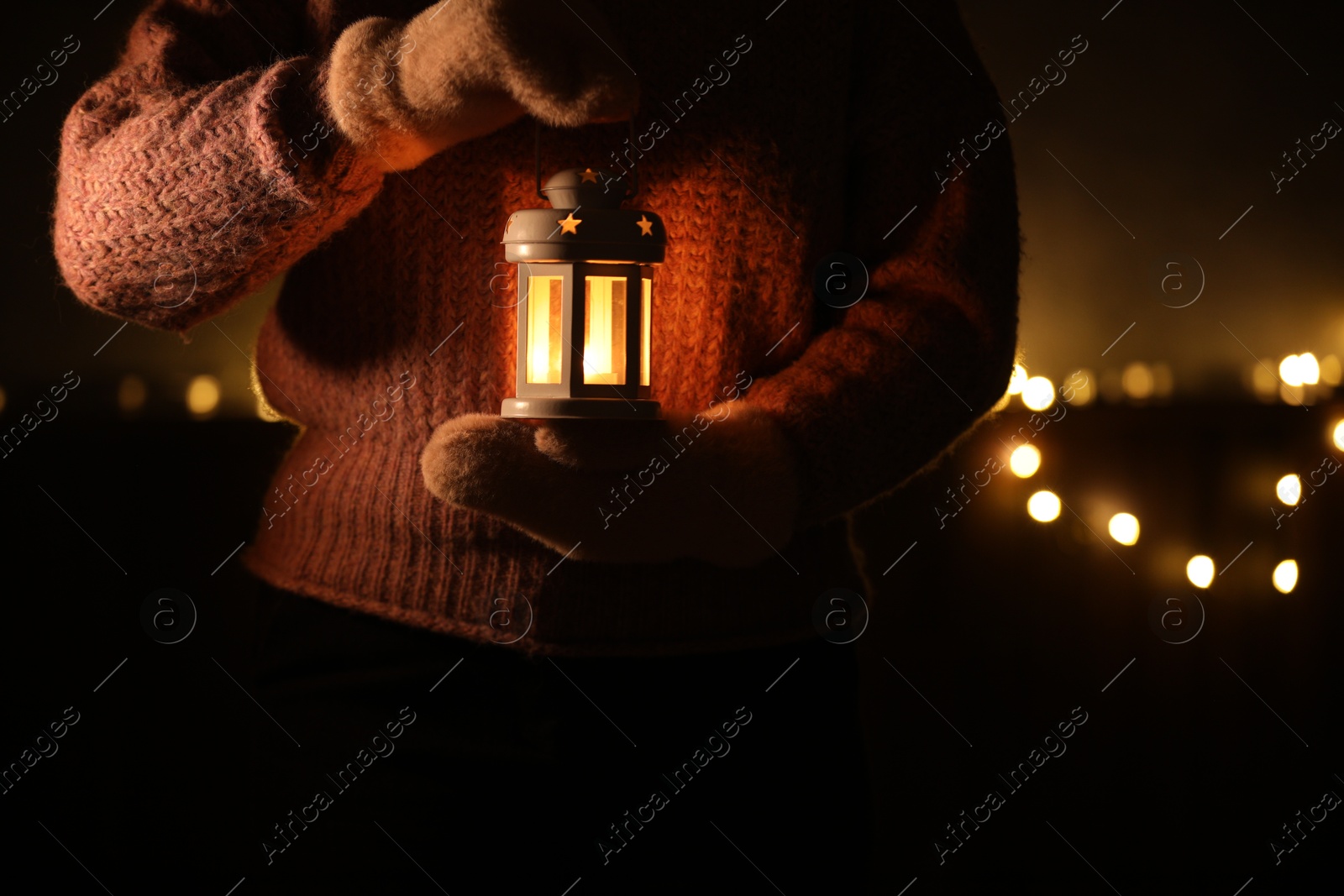 Photo of Woman holding Christmas lantern with burning candle in darkness, closeup. Bokeh effect