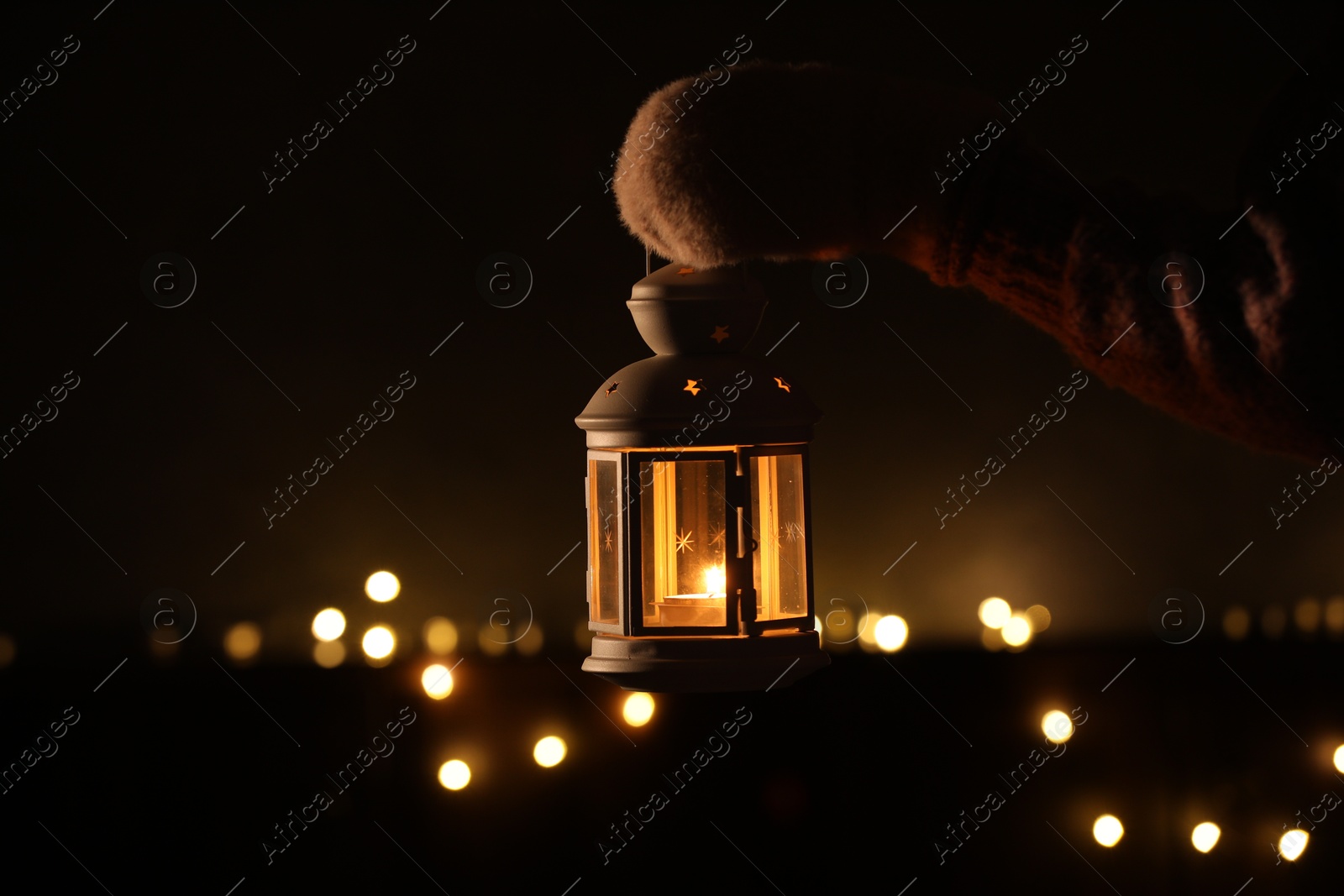 Photo of Woman holding Christmas lantern with burning candle in darkness, closeup. Bokeh effect