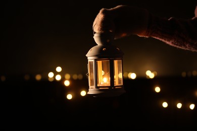 Photo of Woman holding Christmas lantern with burning candle in darkness, closeup. Bokeh effect