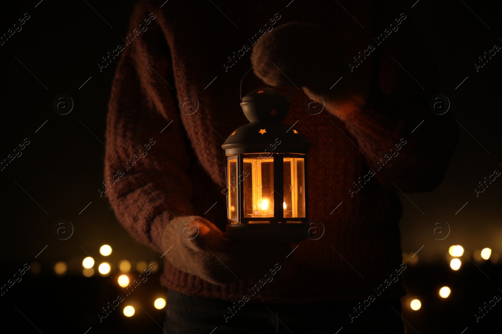 Photo of Woman holding Christmas lantern with burning candle in darkness, closeup. Bokeh effect