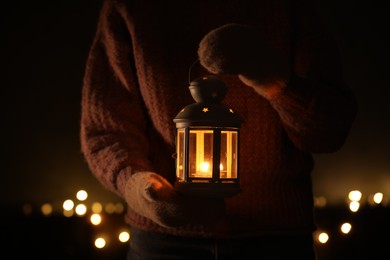 Photo of Woman holding Christmas lantern with burning candle in darkness, closeup. Bokeh effect