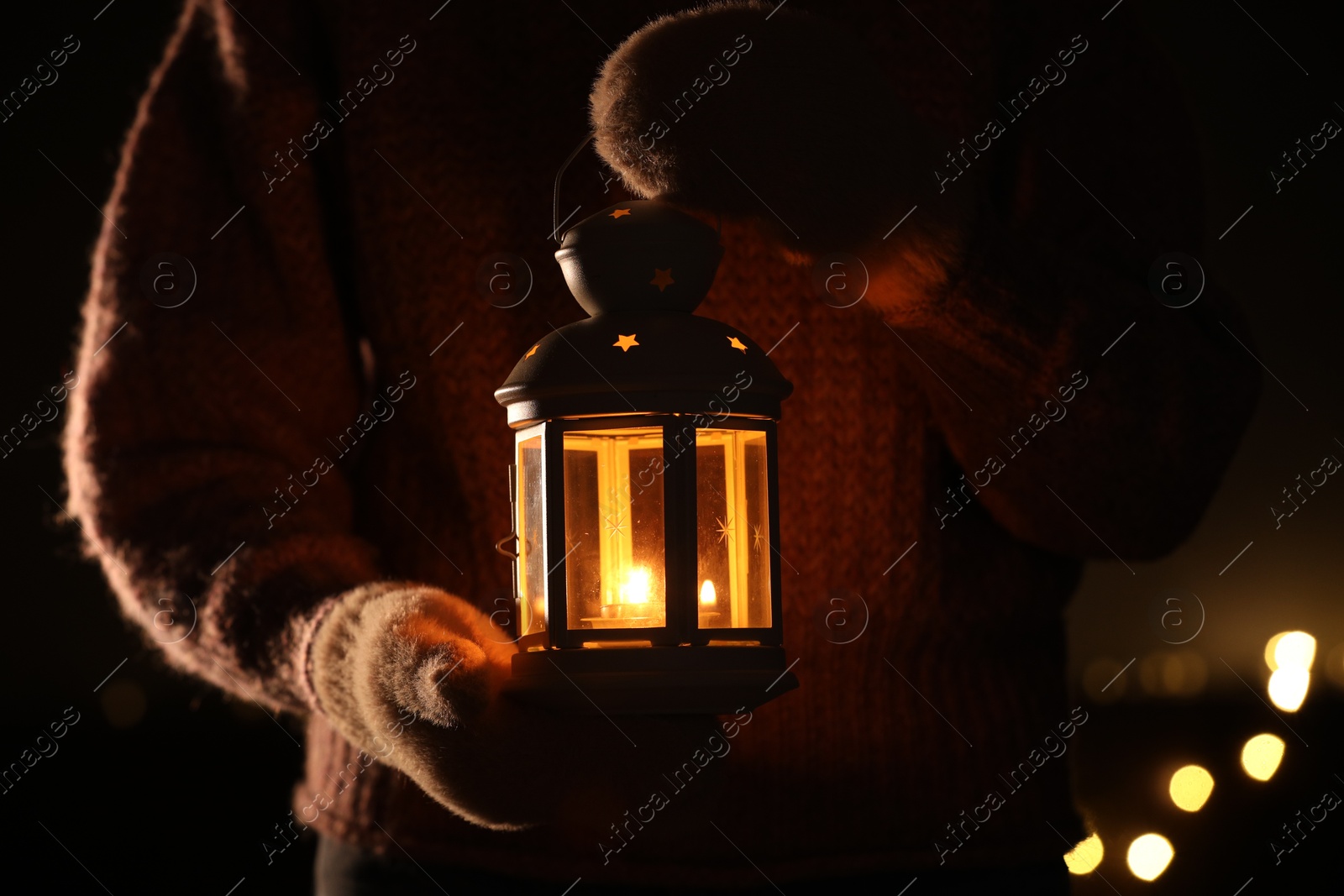 Photo of Woman holding Christmas lantern with burning candle in darkness, closeup. Bokeh effect